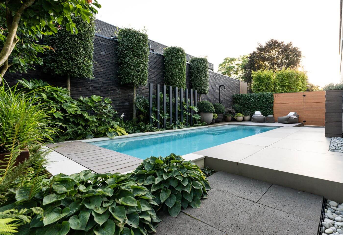 Luxe zwembad in donkere tinten, ge&iuml;ntegreerd in een industri&euml;le stijl tuin met groenblijvende beplanting en een jungle-achtige sfeer.